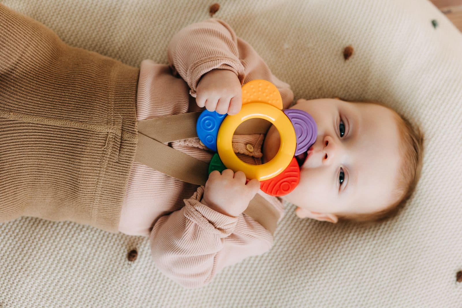 A baby lying on a blanket, chewing on a colored teething ring held in both hands.